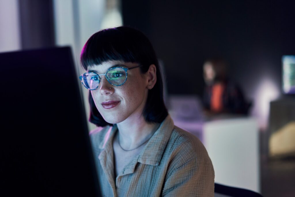 A person with short dark hair and glasses is looking at a computer screen. They are wearing a light-colored shirt and are illuminated by the screen's glow. The background is blurred, showing another person seated in a dimly lit room.