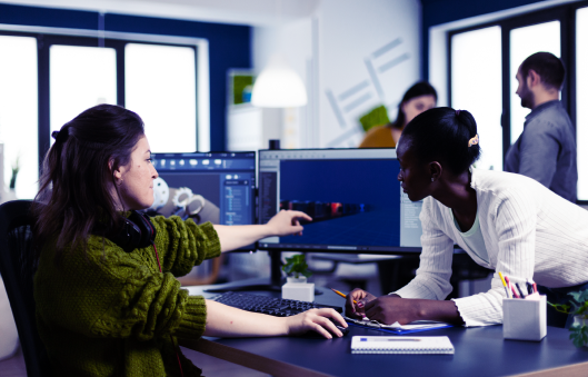 Two women at a desk collaborate on a computer project. One points at a screen displaying a 3D rendering. In the background, a man and woman engage in conversation. The office setting includes multiple monitors and large windows.